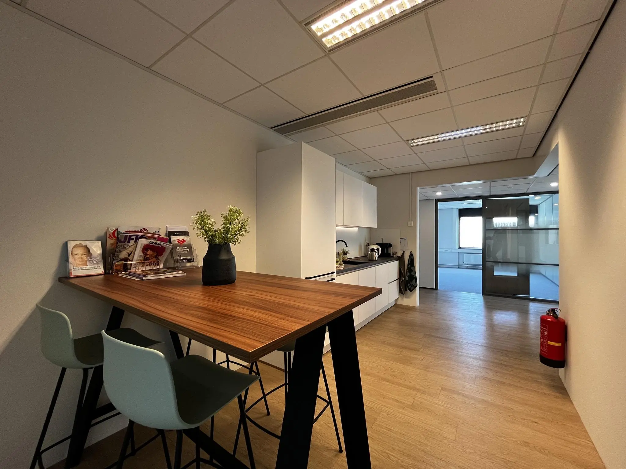 Kleine, moderne kantoorkeuken met hoge tafel, tijdschriftenrek en zicht op vergaderruimte.